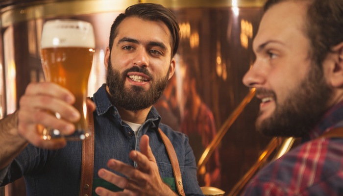 Handsome bearded brewer smiling, talking to his colleague while examining freshly brewed craft beer. Two beermakers working at microbrewery.